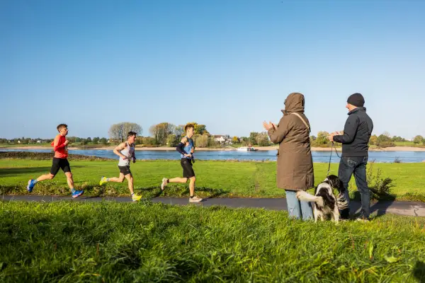 Drei Läufer sind am Rheinufer unterwegs, während Zuschauer sie mit Applaus anfeuern. Die Strecke des Rhein City Run führt über Kaiserswerth und Wittlaer nach Duisburg-Süd. Foto: bunert Events GmbH